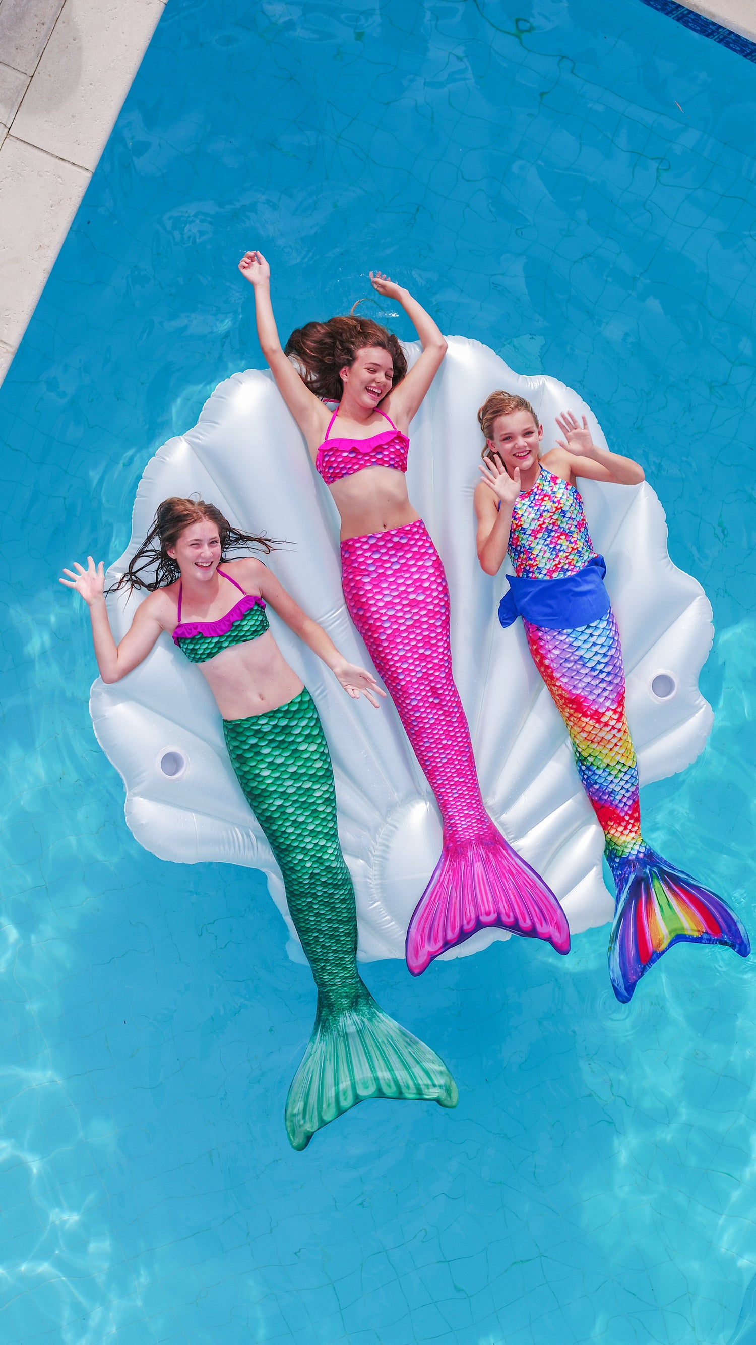 Three children in colorful mermaid tails lying on a seashell-shaped inflatable pool float in a pool.